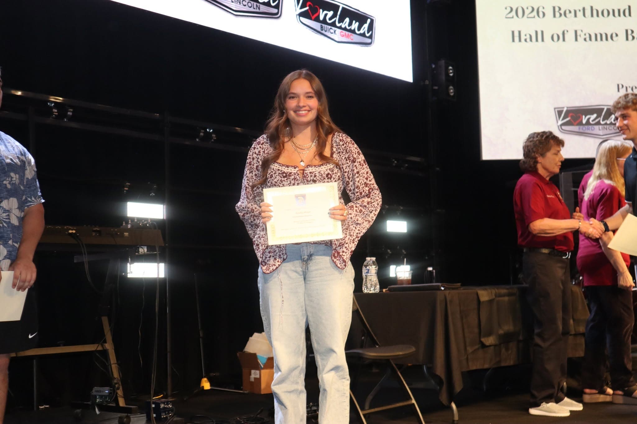 A young woman holding a certificate on stage at a Berthoud Sports Hall of Fame event